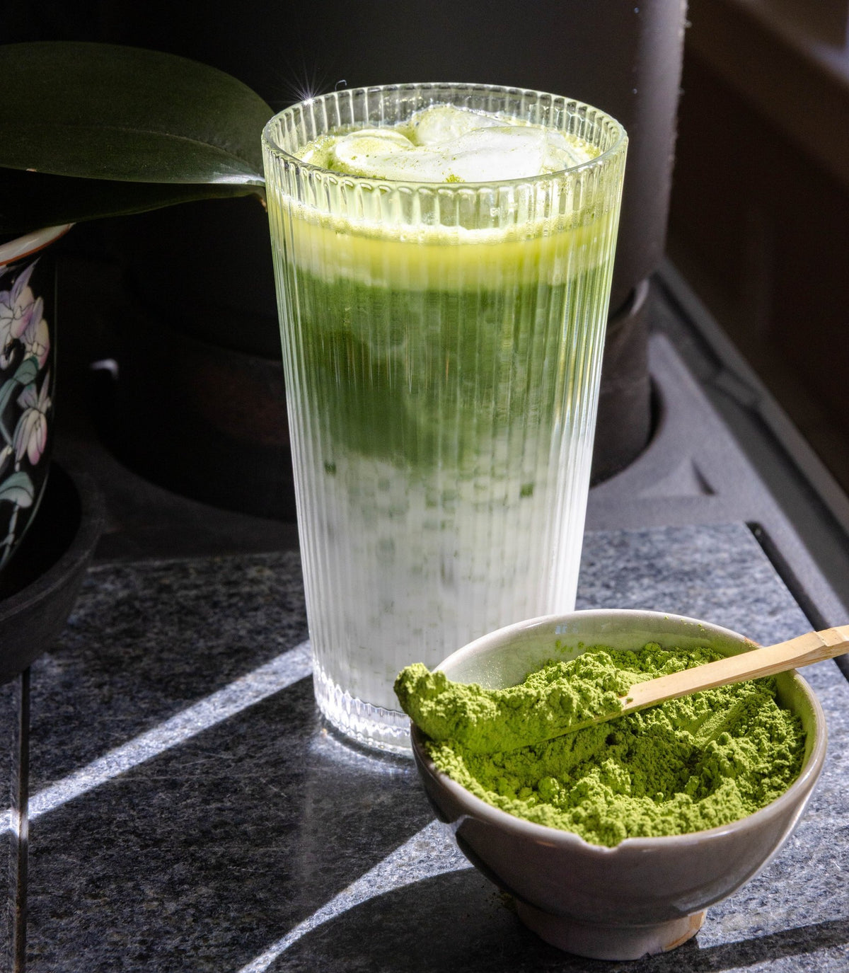 A layered iced matcha latte in a ribbed glass beside a bowl of matcha powder and a bamboo scoop.