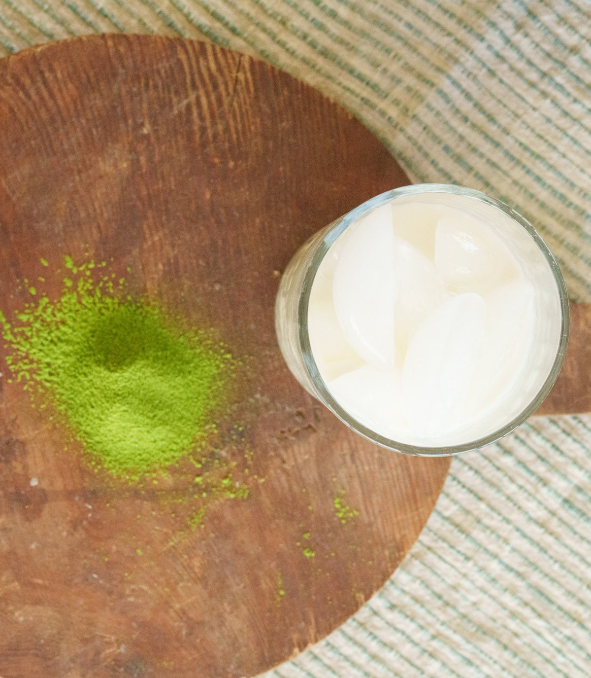 Matcha powder sifted onto a round wooden board beside a glass of iced milk, seen from above.