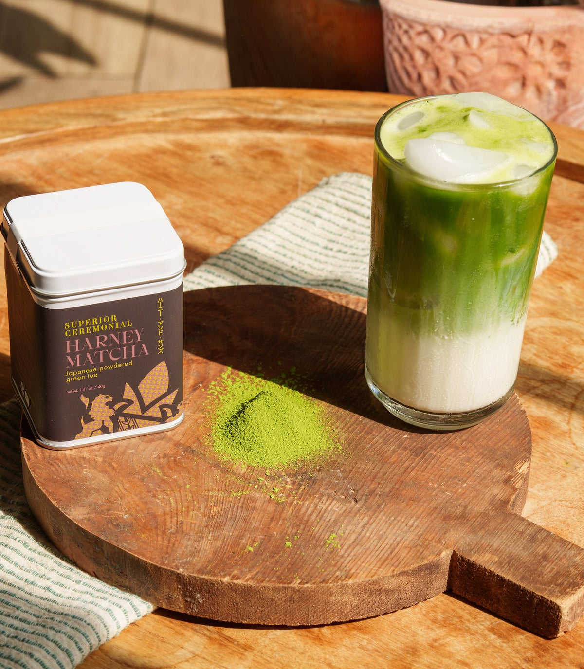 A Superior Ceremonial Matcha tin, a mound of matcha powder, and a layered iced matcha latte on a round wooden board in sunlight.