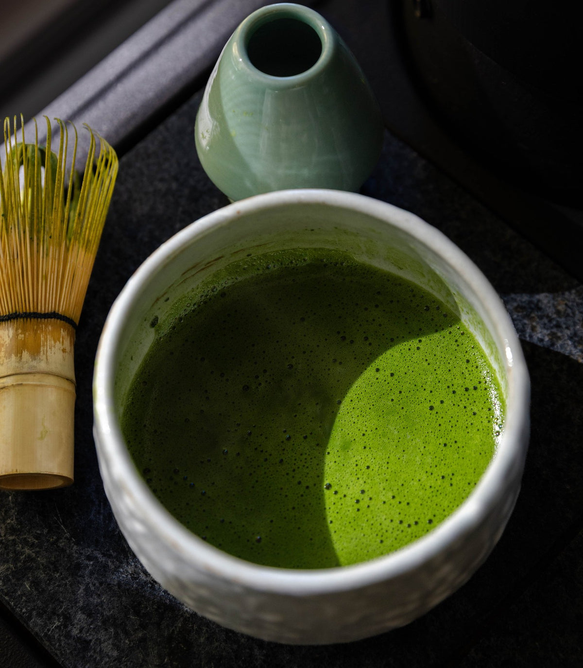 Whisked matcha in a white bowl beside a bamboo whisk and whisk stand, in bright sunlight.