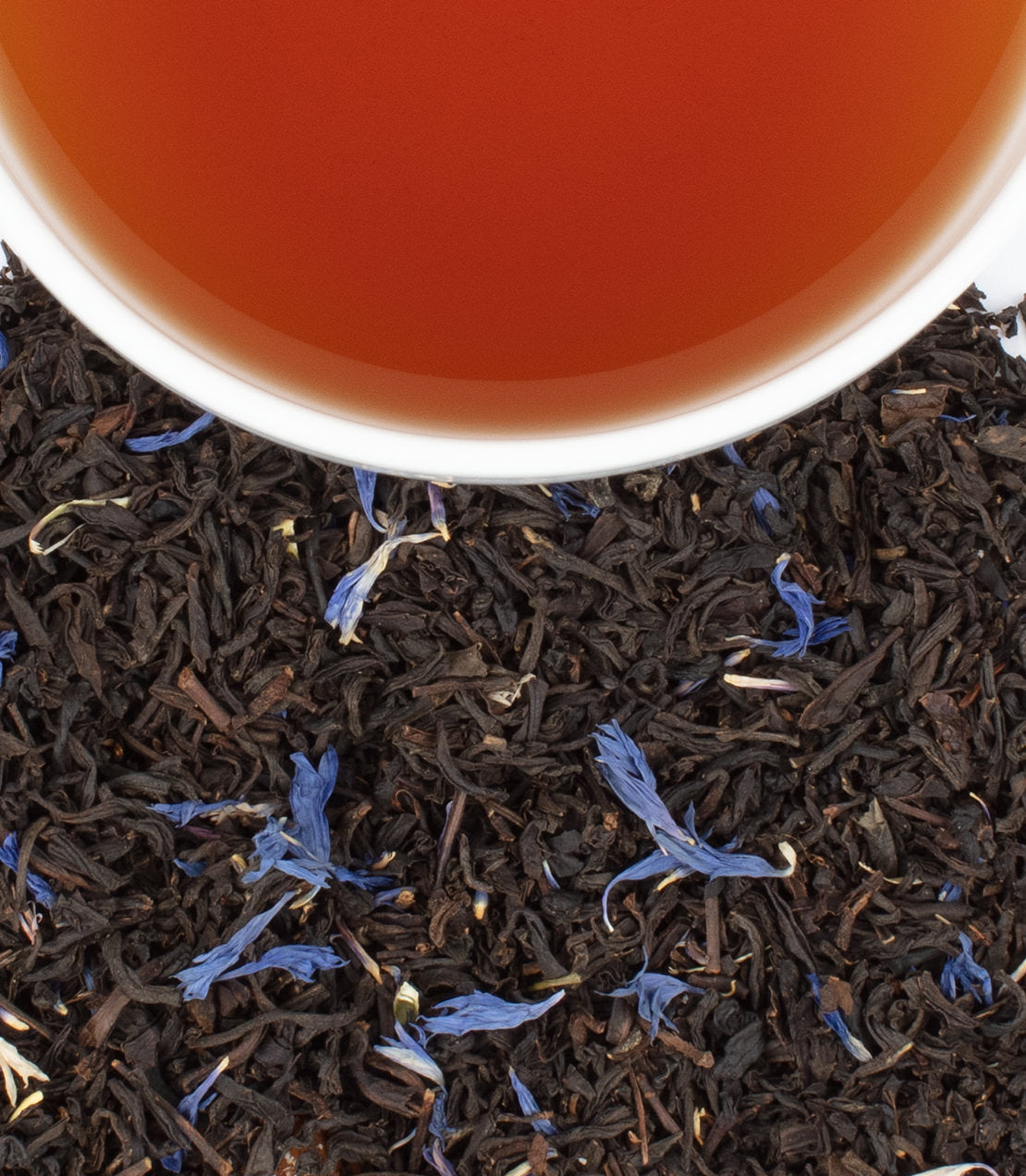 A cup of brewed tea sits atop loose black tea leaves and blue flower petals, showcasing a celebration-themed tin of 30 sachets.