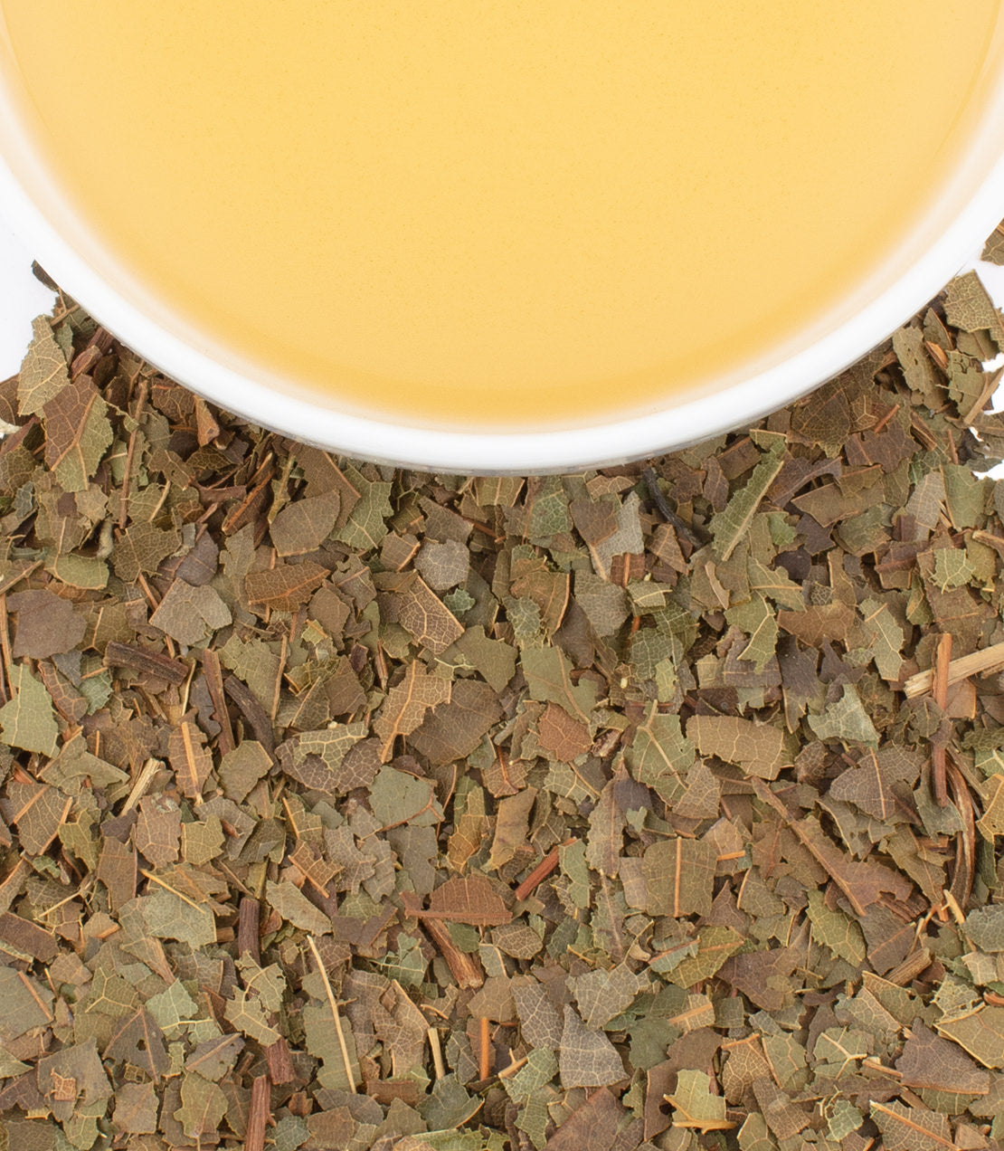 Dried avocado leaves scattered around a white bowl filled with light yellow avocado leaf tea.