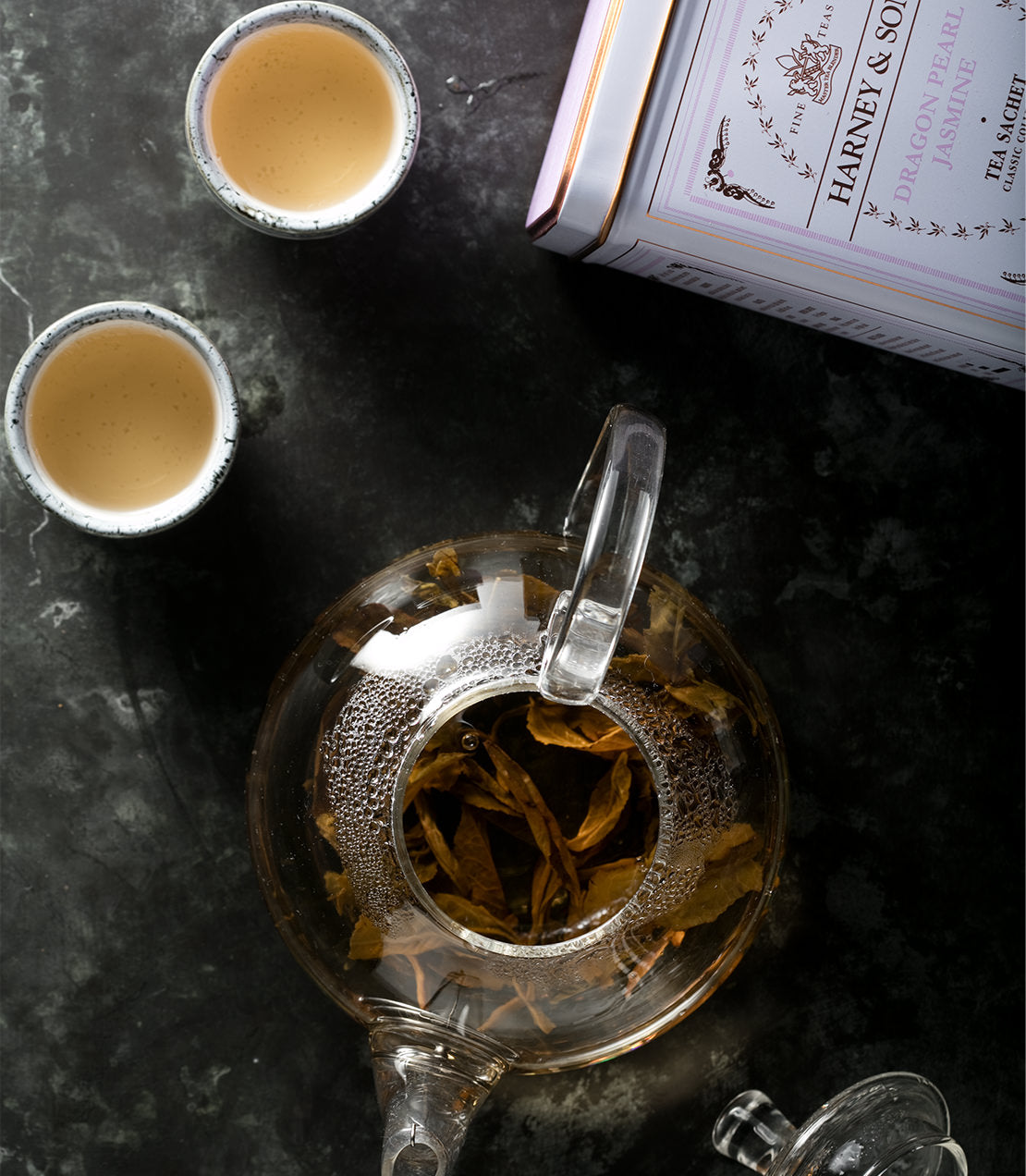 Classic tin of Dragon Pearl Jasmine tea sachets with brewed tea in a glass teapot and two cups.