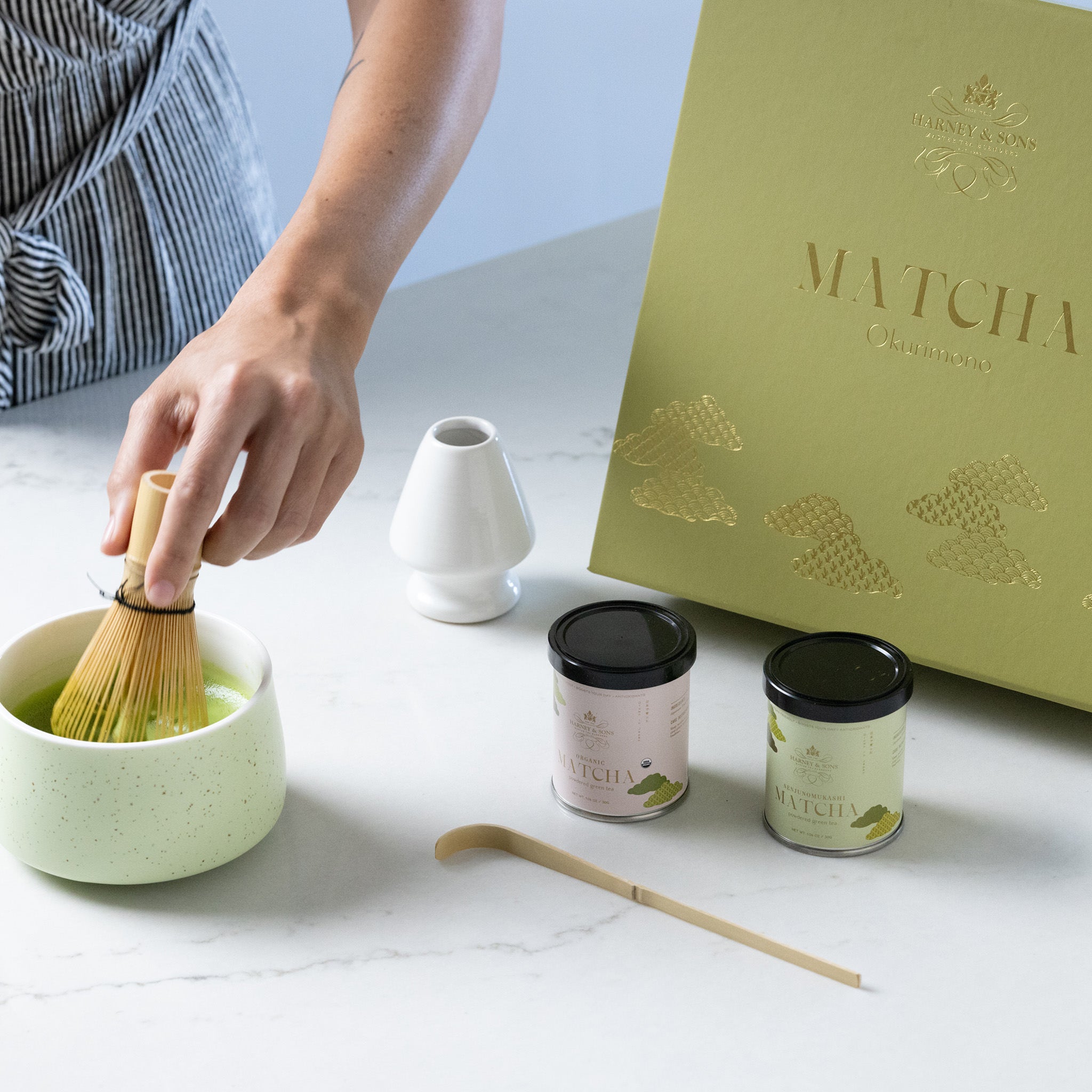 Hand whisking matcha in a bowl with gift box and canisters of matcha in the background.