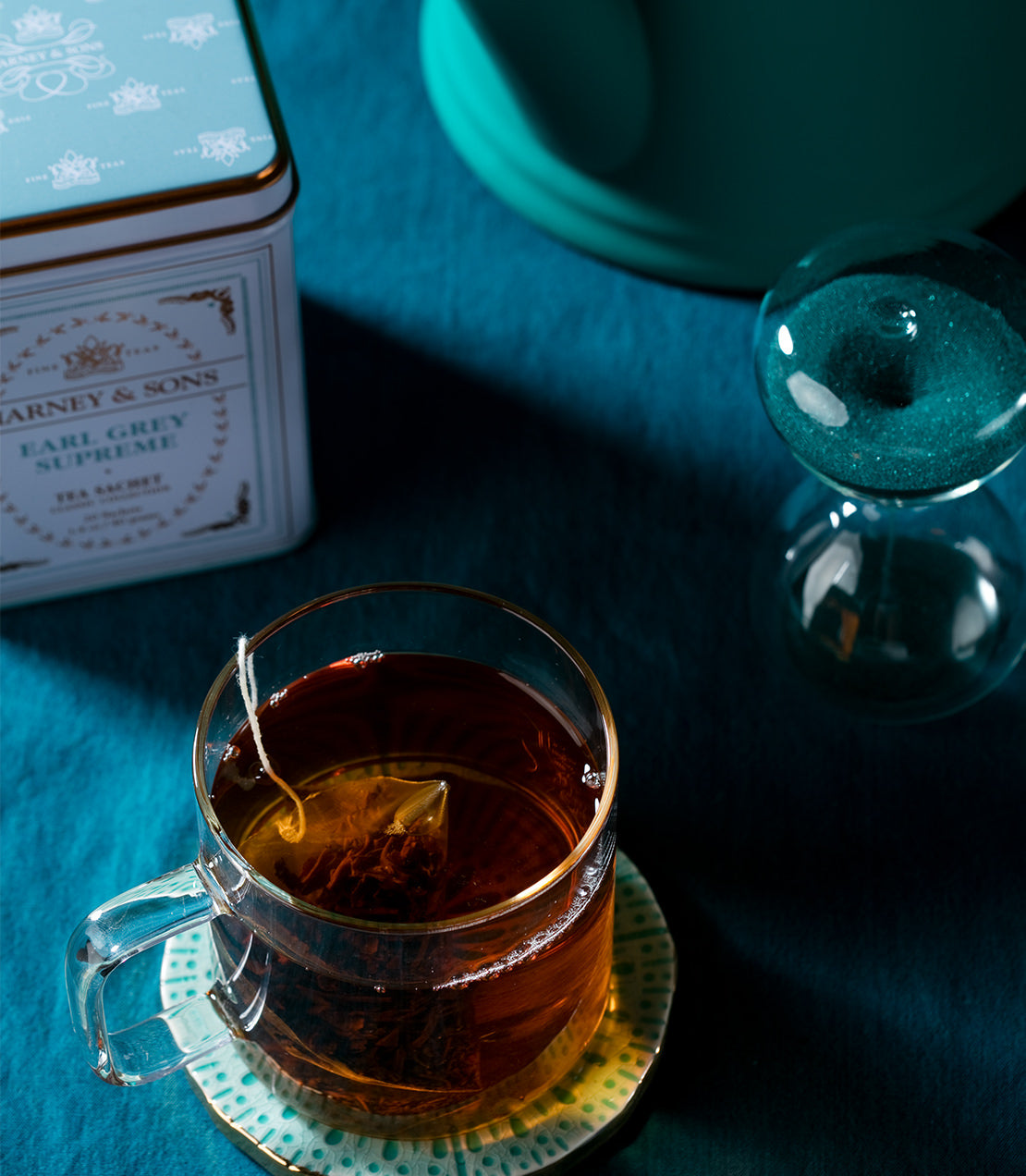 A glass cup of Earl Grey Supreme tea with a tea sachet, beside a classic tin and an hourglass on a teal background.