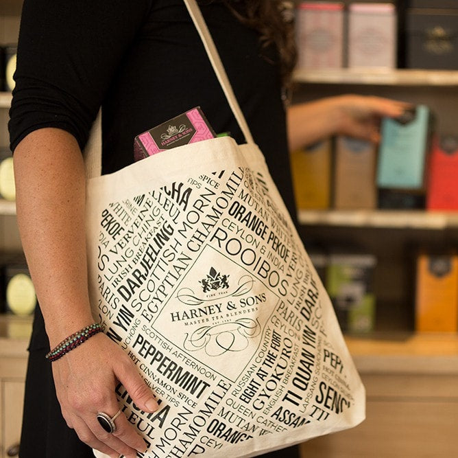Harney & Sons tote bag featuring tea-themed designs, held by a person in a store with colorful tea boxes.